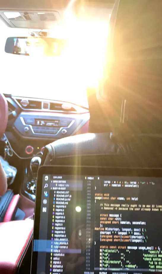 Inside a car at sunset, a person drives with the steering wheel and dashboard visible, while a laptop displaying code rests on the back seat.