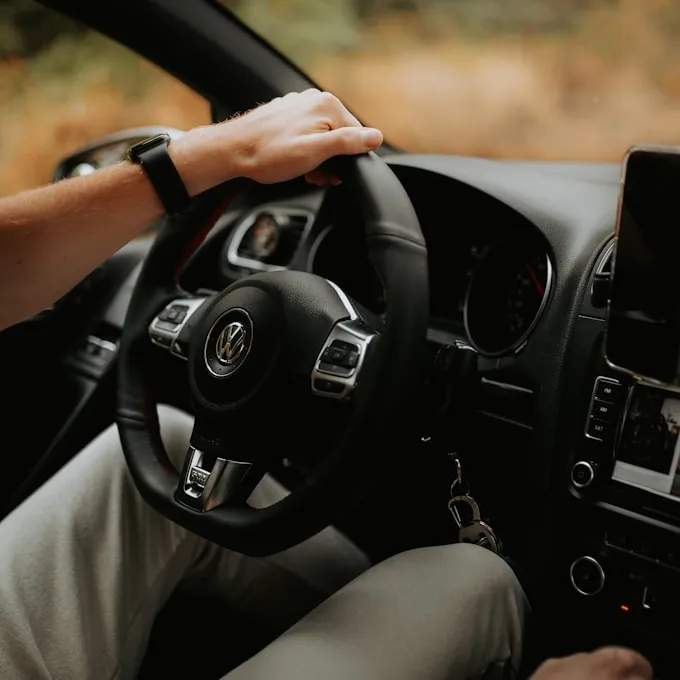 A person sitting in the driver seat of a car holding the steering wheel, with dashboard controls and a mounted screen visible.