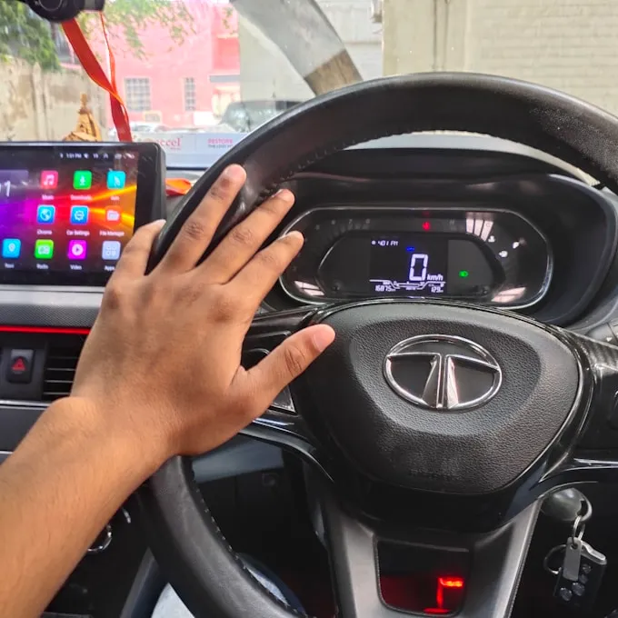 A hand resting on the steering wheel of a car with a touchscreen dashboard display and a blurred cityscape beyond the windscreen.